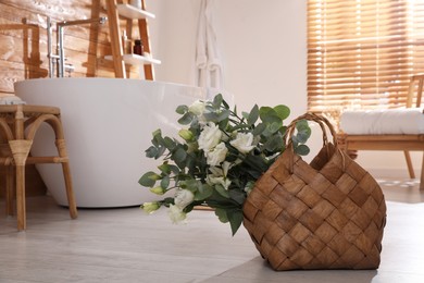 Stylish wicker basket with fresh eucalyptus branches and flowers on floor in bathroom