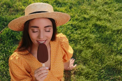 Photo of Beautiful young woman eating ice cream glazed in chocolate on green grass outdoors. Space for text