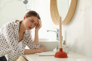 Photo of Unhappy young woman with plunger near clogged sink in bathroom