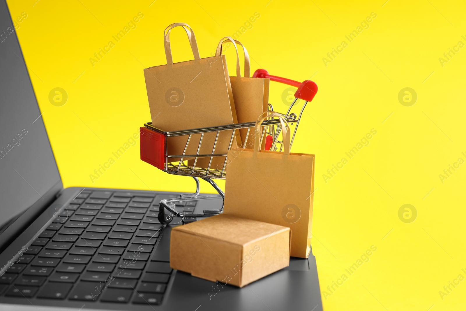 Photo of Online store. Laptop, mini shopping cart and purchases on yellow background, closeup