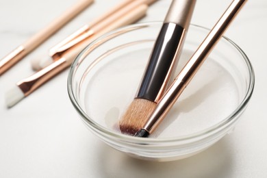 Photo of Clean makeup brushes in bowl with cleanser on white table, closeup