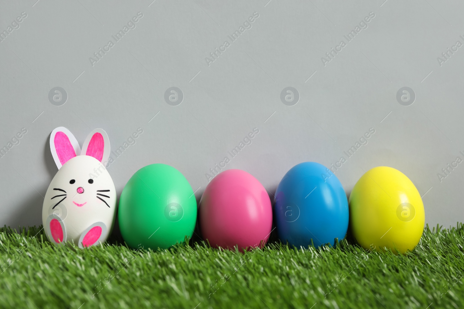 Photo of Bright eggs and white one as Easter bunny on green grass against grey background