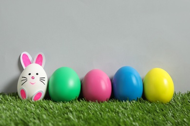 Bright eggs and white one as Easter bunny on green grass against grey background