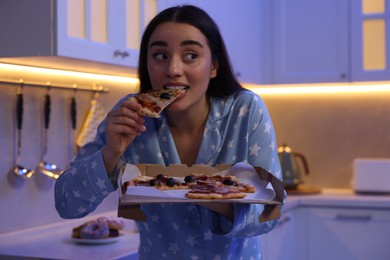 Photo of Young woman eating pizza in kitchen at night. Bad habit