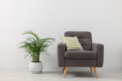 Photo of Comfortable armchair with cushion and houseplant near white wall indoors. Interior design
