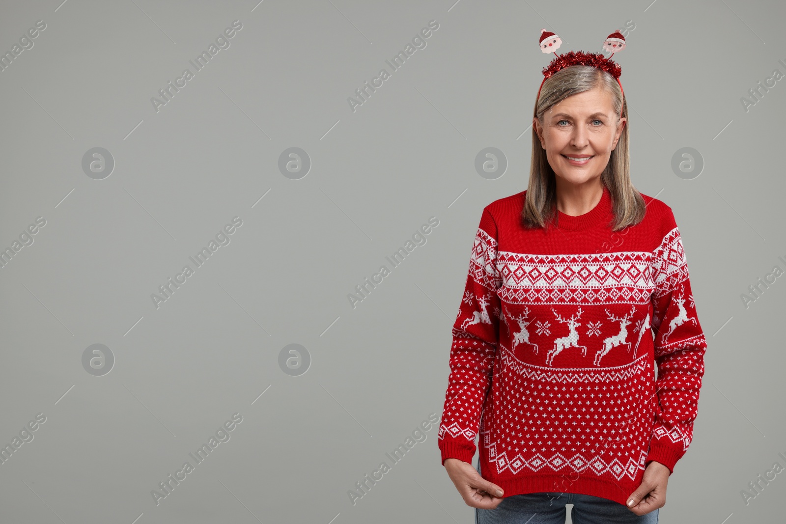 Photo of Happy senior woman in Santa headband showing her Christmas sweater on grey background. Space for text