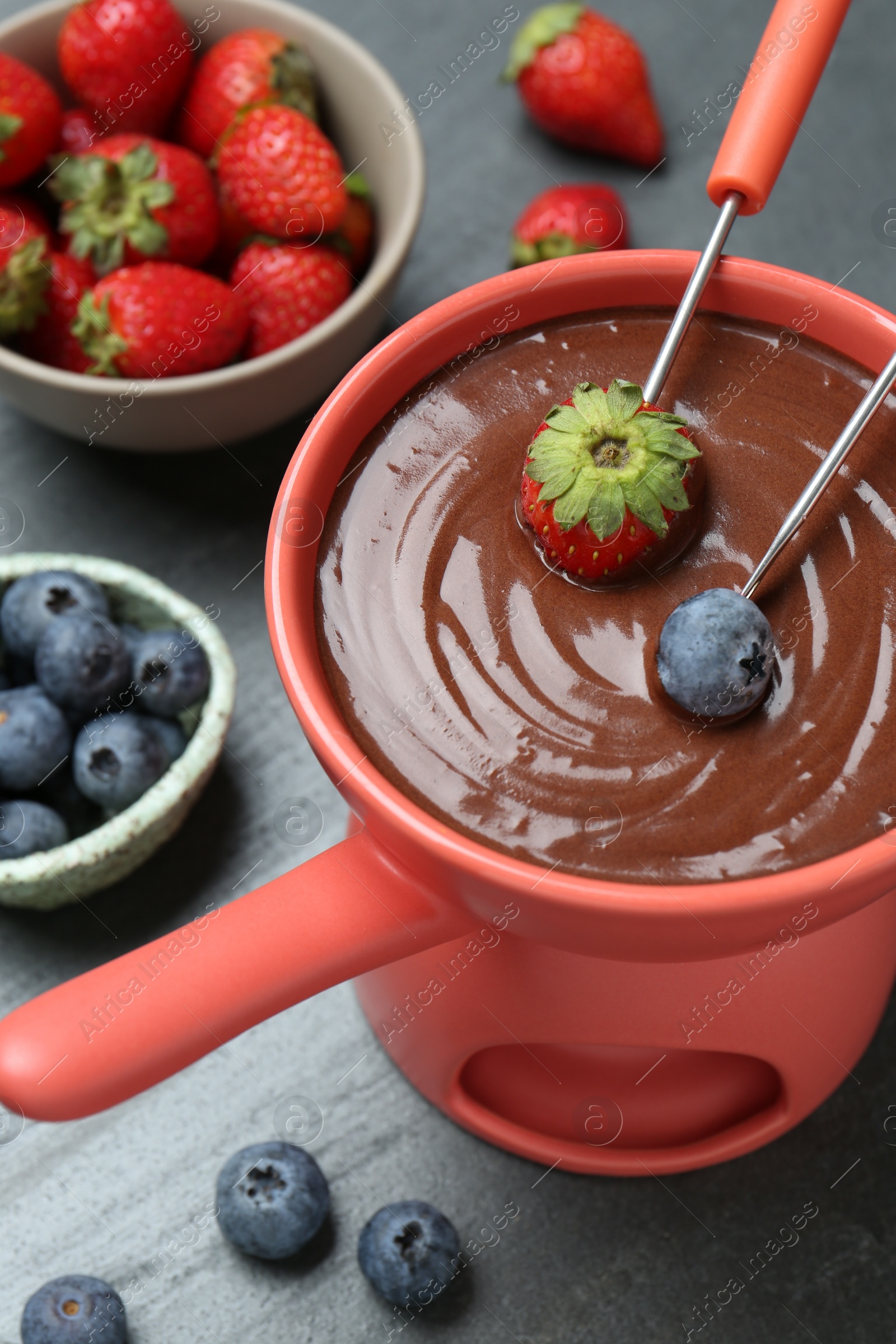 Photo of Dipping fresh berries in fondue pot with melted chocolate at grey table, above view