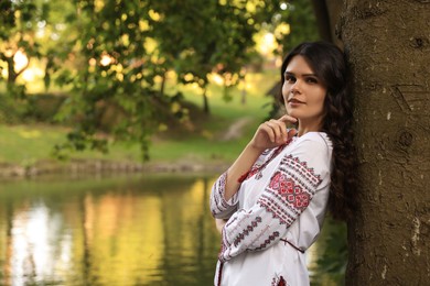 Photo of Beautiful woman in embroidered shirt near lake, space for text. Ukrainian national clothes