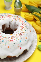 Photo of Easter cake with sprinkles, painted eggs and tulips on yellow background, closeup