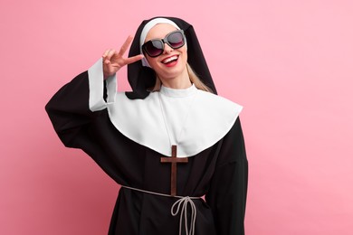Photo of Happy woman in nun habit and sunglasses showing V-sign against pink background