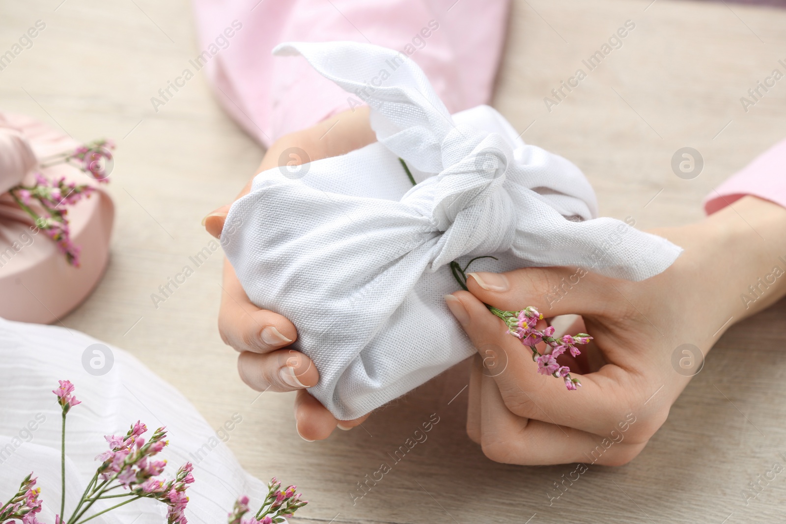 Photo of Furoshiki technique. Woman decorating gift wrapped in white fabric with beautiful pink flower at wooden table, closeup