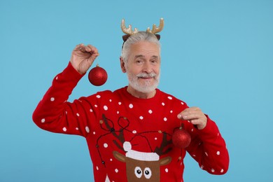 Photo of Senior man in Christmas sweater and reindeer headband holding festive baubles on light blue background