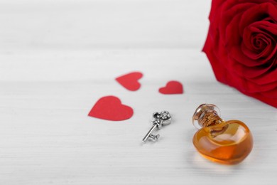 Heart shaped bottle of love potion with small key, paper hearts and red rose on white wooden table, space for text