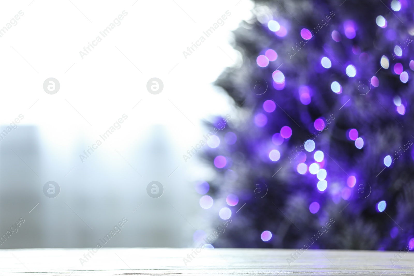 Photo of Blurred view of beautiful Christmas tree with purple lights near window indoors, focus on wooden table. Space for text