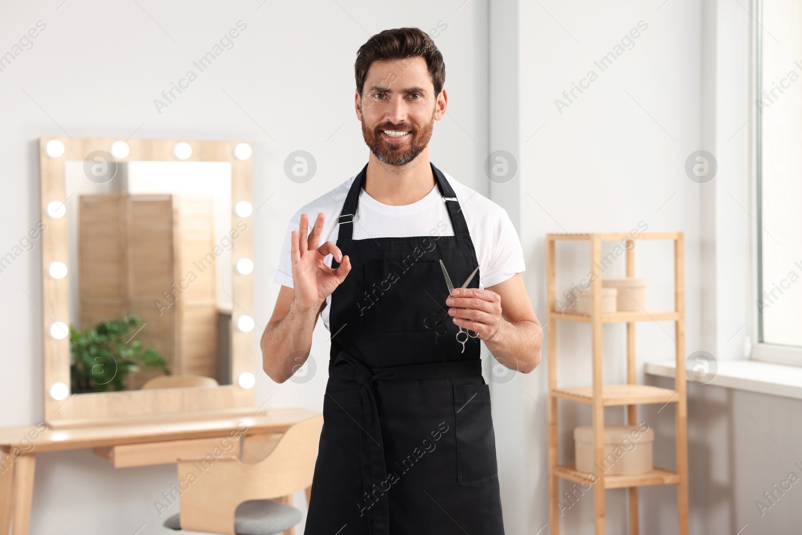 Photo of Smiling hairdresser in apron holding scissors and showing ok gesture near vanity mirror in salon