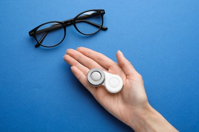 Photo of Woman holding case with contact lenses near glasses on blue background, top view