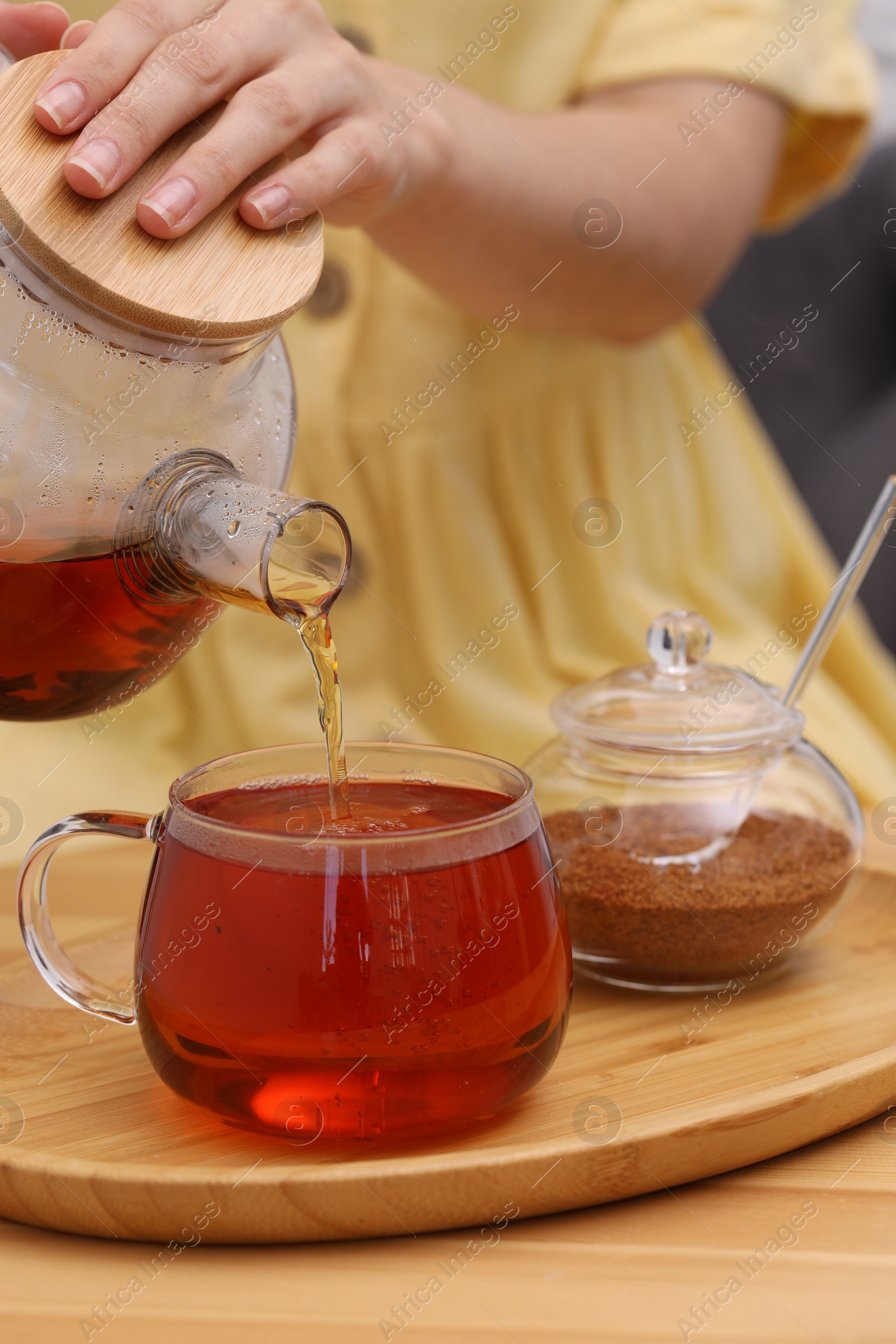 Photo of Woman pouring aromatic hot tea into glass cup at wooden table, closeup