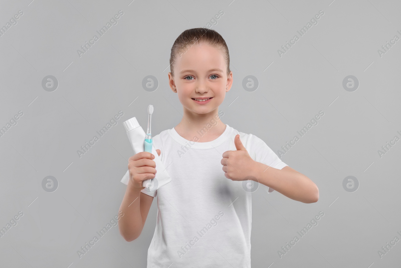 Photo of Happy girl holding electric toothbrush with tube of toothpaste and showing thumb up on light grey background