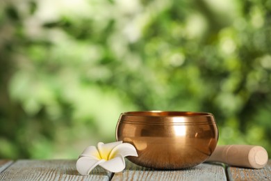 Photo of Golden singing bowl, mallet and flower on wooden table outdoors