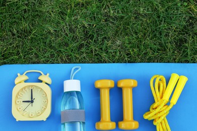 Photo of Alarm clock, skipping rope, dumbbells, bottle of water and fitness mat on green grass, flat lay with space for text. Morning exercise