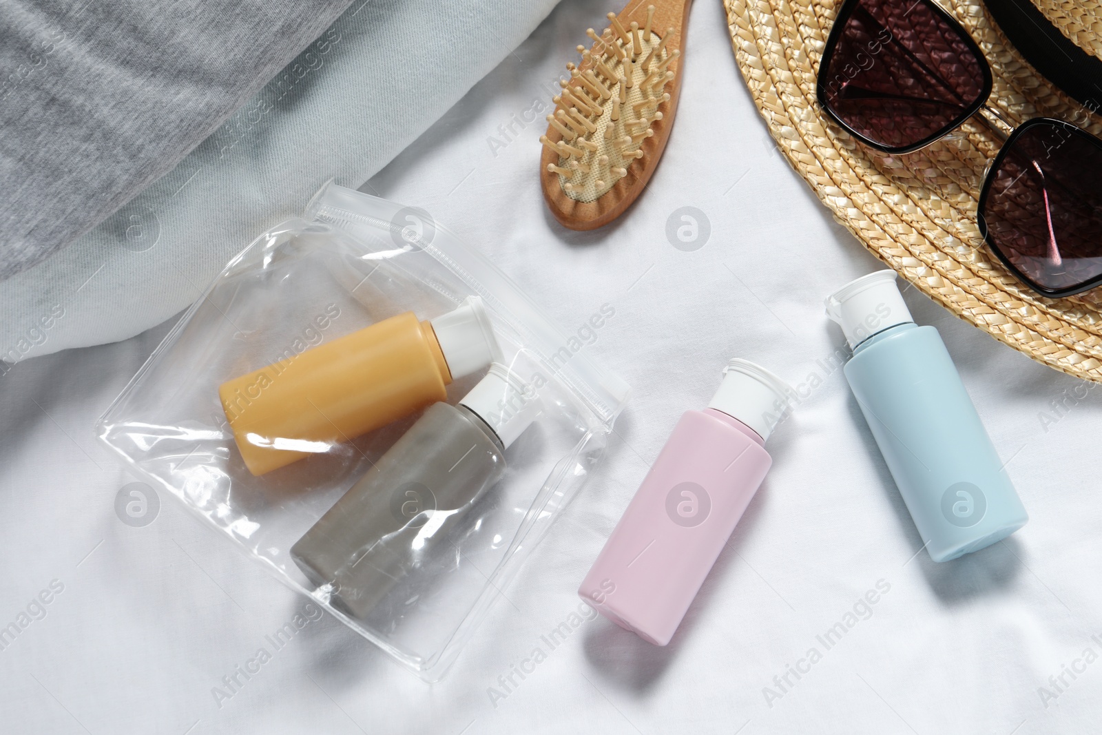 Photo of Cosmetic travel kit. Plastic bag with small containers of personal care products, beach accessories and brush on bed, flat lay