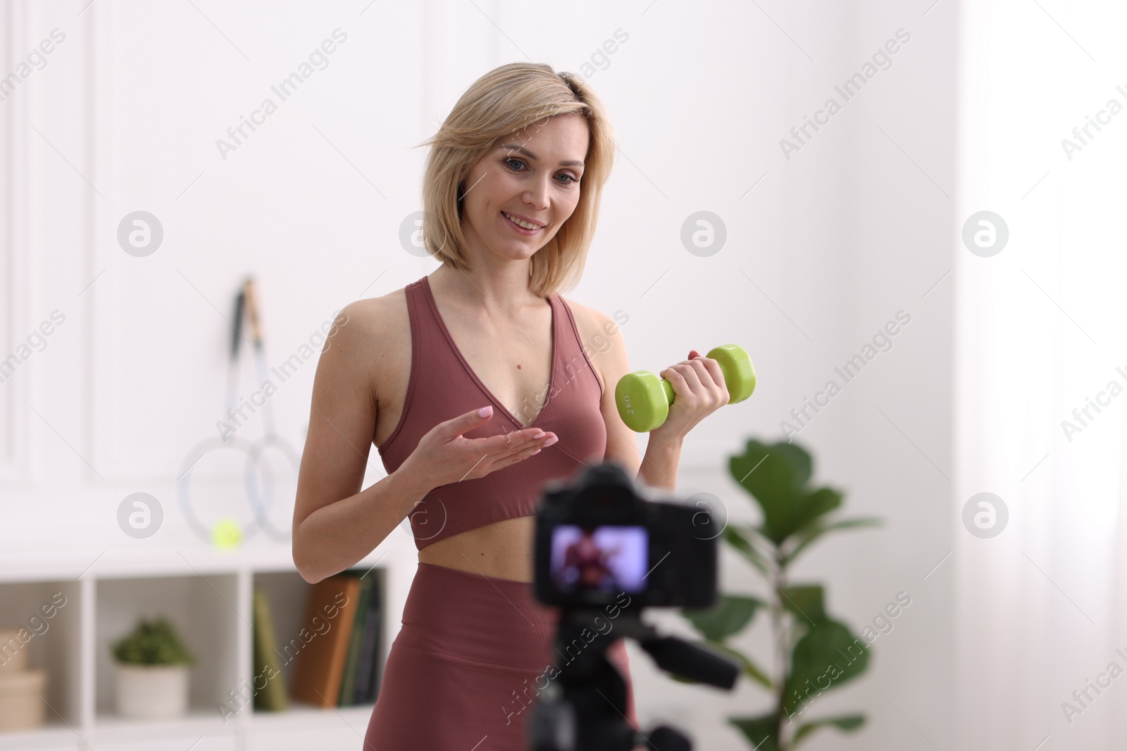 Photo of Smiling sports blogger with dumbbell recording fitness lesson with camera at home