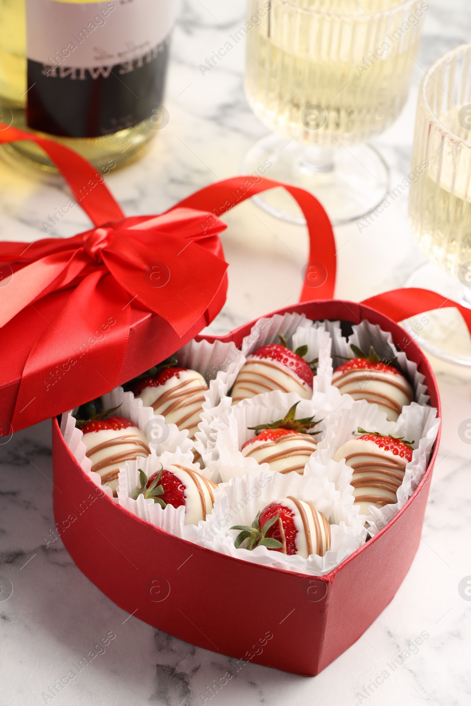 Photo of Delicious chocolate covered strawberries in heart shaped box and sparkling wine on white marble table