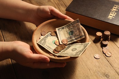 Photo of Donate and give concept. Woman holding bowl with money at wooden table, closeup