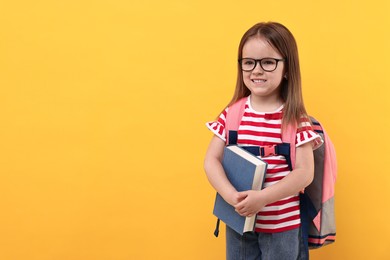 Photo of Cute little girl in glasses with book and backpack on orange background. Space for text