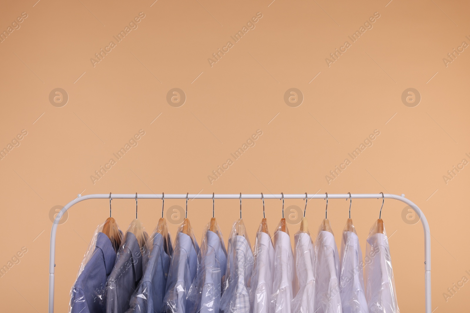 Photo of Dry-cleaning service. Many different clothes in plastic bags hanging on rack against beige background, space for text