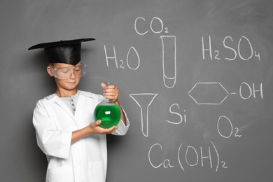 Little school child in laboratory uniform with flask of liquid and chemical formulas on grey background