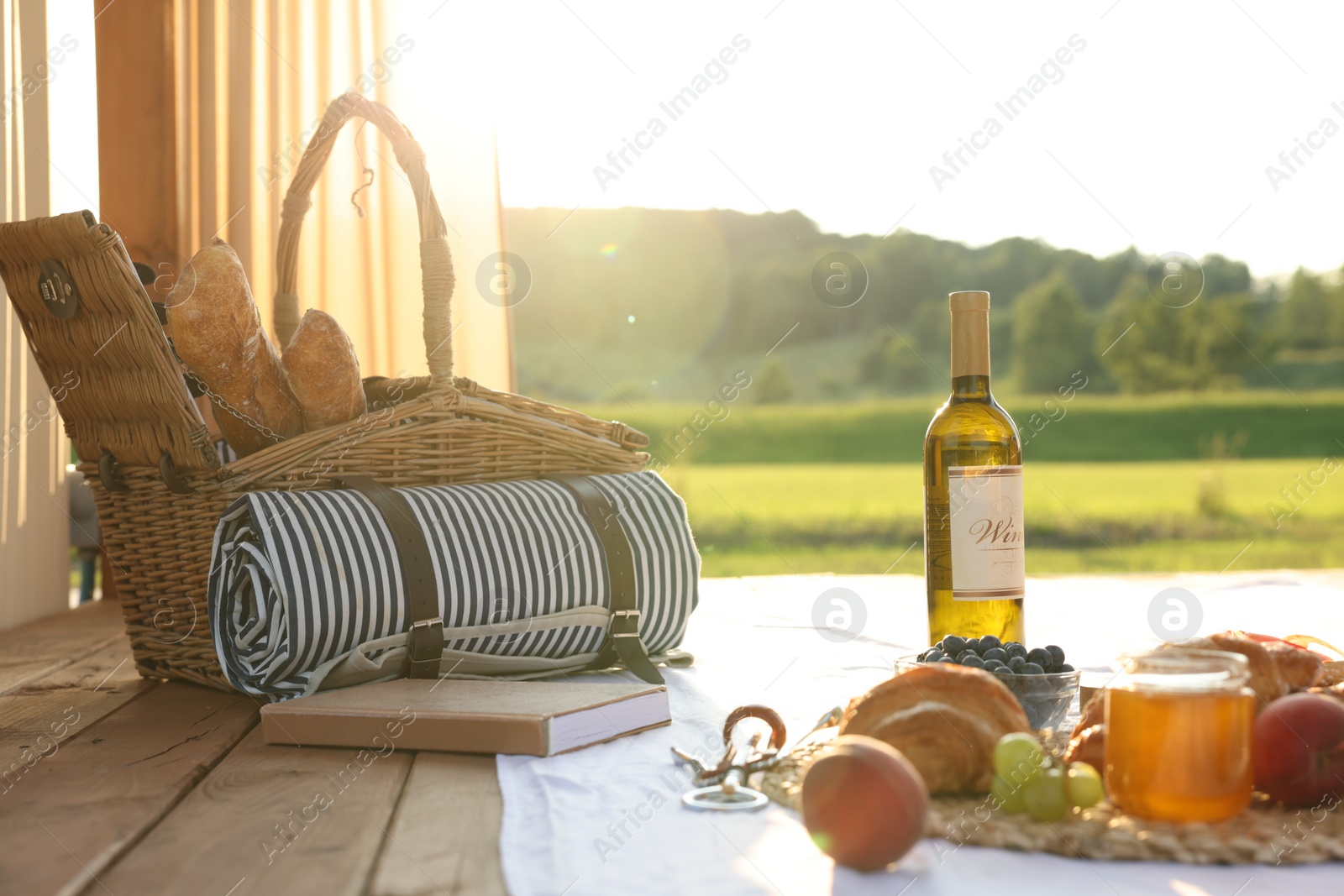 Photo of Romantic date. Wicker basket, glass of wine and snacks for picnic on white blanket outdoors