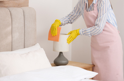 Young chambermaid wiping dust from lamp in bedroom, closeup