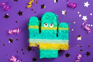 Photo of Cactus shaped pinata, streamers and glitter on purple background, flat lay