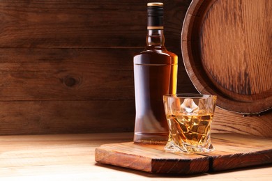 Photo of Whiskey with ice cubes in glass, bottle and barrel on wooden table, space for text