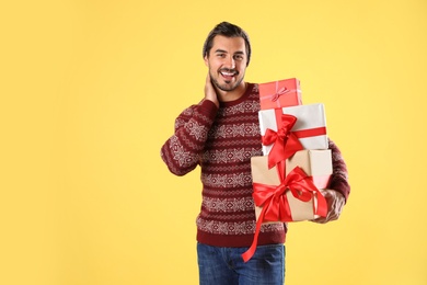 Happy man in Christmas sweater and hat holding gift boxes on yellow background