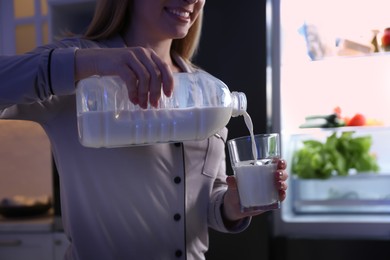 Young woman pouring milk from gallon bottle into glass near refrigerator in kitchen at night, closeup