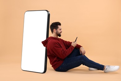 Image of Man with mobile phone sitting near huge device with empty screen on dark beige background. Mockup for design