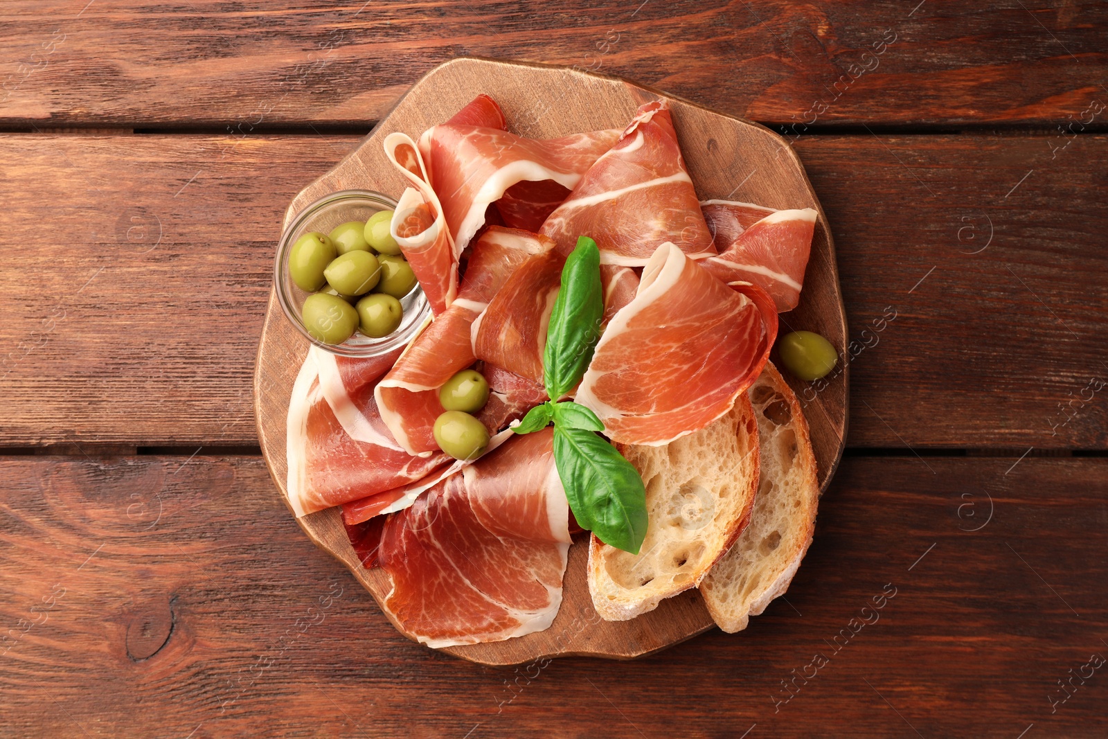 Photo of Slices of tasty cured ham, olives, bread and basil on wooden table, top view