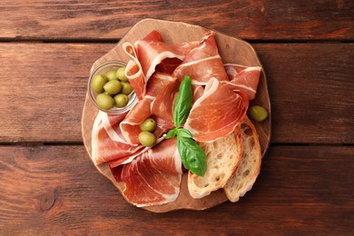 Photo of Slices of tasty cured ham, olives, bread and basil on wooden table, top view