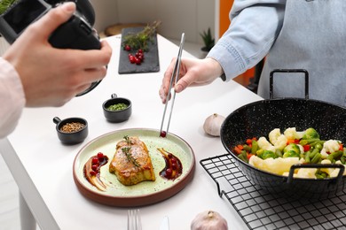 Professional photographer taking photo while food stylist decorating composition with meat medallion in studio, closeup