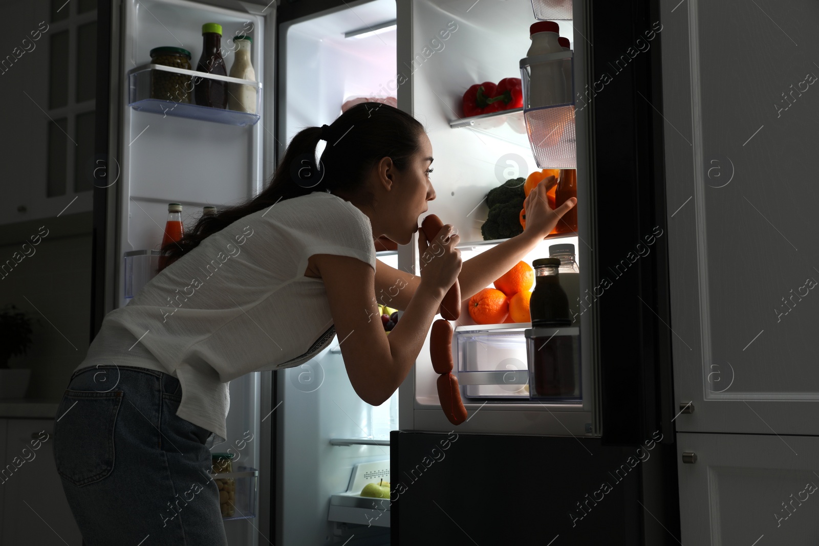 Photo of Young woman eating sausages near open refrigerator at night