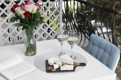 Vase with roses, open book, glasses of wine and snacks on white table at balcony