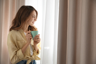 Photo of Woman holding cup near window with beautiful curtains at home