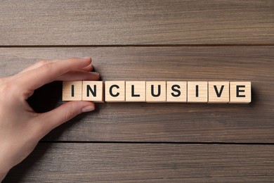 Photo of Woman making word Inclusive with cubes on wooden table, closeup with top view