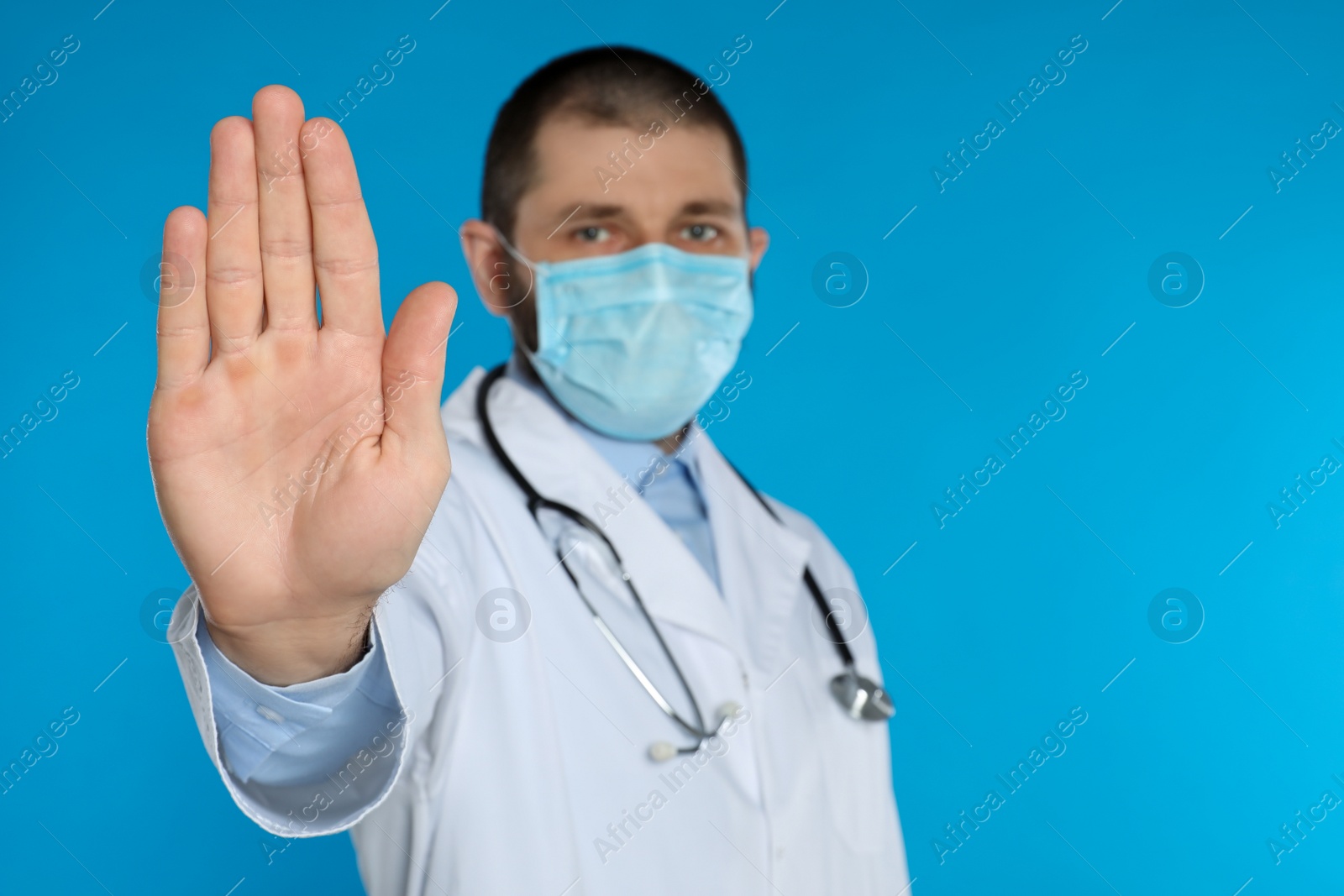 Photo of Doctor in protective mask showing stop gesture on light blue background, space for text. Prevent spreading of coronavirus