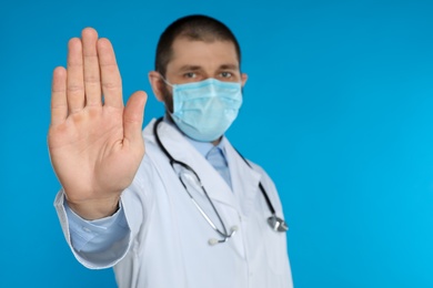 Photo of Doctor in protective mask showing stop gesture on light blue background, space for text. Prevent spreading of coronavirus