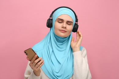 Muslim woman in hijab and headphones listening to music with smartphone on pink background