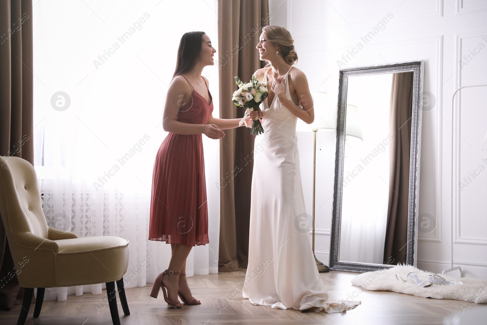 Photo of Gorgeous bride in beautiful wedding dress and her friend near window in room