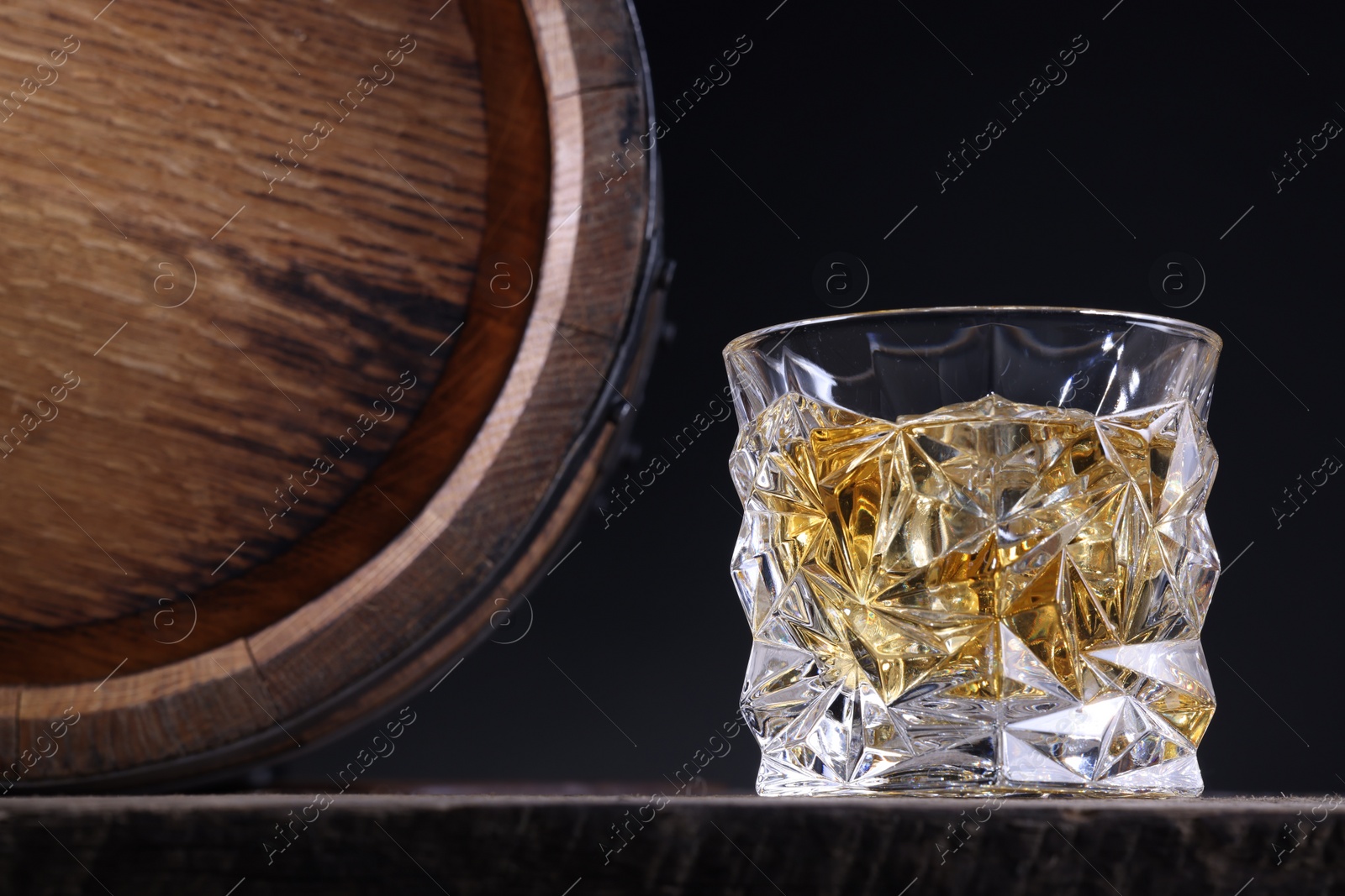 Photo of Whiskey with ice cubes in glass and barrel on wooden table against black background, closeup. Space for text
